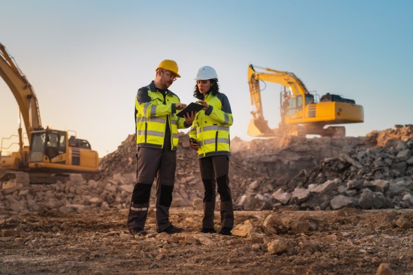 Caucasian Male Civil Engineer Talking To Hispanic Female Inspector And Using Tablet On Construction Site of New Apartment Complex. Real Estate Developers Discussing Business, Excavators Working.